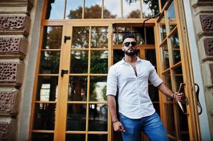 Stylish tall arabian man model in white shirt, jeans and sunglasses posed at street of city. Beard attractive arab guy against entrance door of restaurant. photo