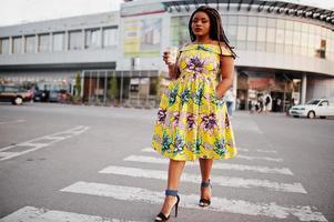 Cute small height african american girl with dreadlocks, wear at coloured yellow dress, walking at crosswalk against trade center with cup of coffee at hand. photo