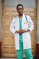 elegante médico afroamericano con estetoscopio y bata de laboratorio contra la puerta del hospital. foto