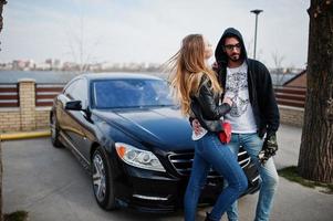 una pareja multirracial genial se abraza contra un elegante auto deportivo negro. foto