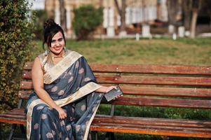 Elegant brunette south asian indian girl in saree sitting on bench with mobile phone at hands. photo