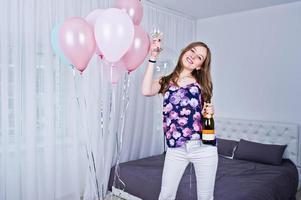 Happy girl with colored balloons on bed at room with glasses and bottle of champagne. Celebrating birthday theme. photo