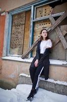 Fashionable long legs brunette model in long black cloak posed outdoor at winter day against old grunge wall with broken windows. photo