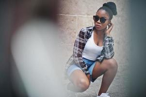 Hip hop african american girl on sunglasses and jeans shorts. Casual street fashion portrait of black woman. photo