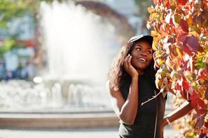 Stylish african american girl in cap posed at sunny autumn day against red leaves. Africa model woman. photo