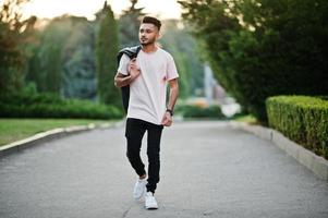 Stylish indian beard man at pink t-shirt with black leather jacket on the shoulder. India model walking at evening city. photo