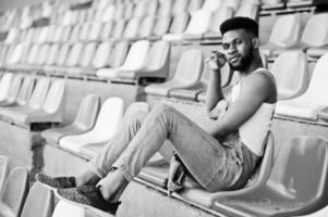 Handsome african american man at jeans overalls posed on colored chairs at stadium. Fashionable black man portrait. photo