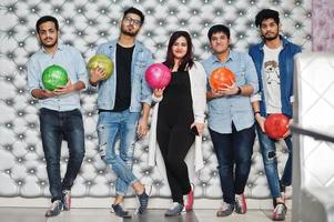 Group of five south asian peoples having rest and fun at bowling club, posing against silver wall with balls at hands. photo