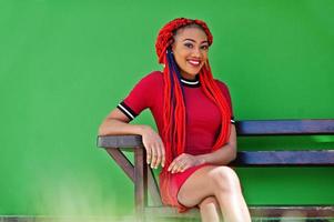 Cute and slim african american girl in red dress with dreadlocks posed on bench background green wall. Stylish black model. photo