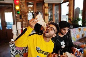 Asian friends guys eating pizza during party at pizzeria. Happy indian people having fun together, eating italian food and sitting on couch. photo