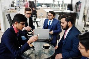 grupo de cinco hombres de negocios indios en trajes sentados en la oficina en el café mirando la computadora portátil y tomando café. foto