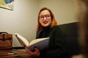 Cheerful young beautiful redhaired woman in glasses, wear on green warm wool sweater,  sitting at cafe and using her notebook. photo