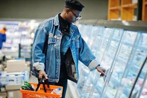 Stylish casual african american man at jeans jacket and black beret holding basket, standing near gastronomic fridge and shopping at supermarket. photo