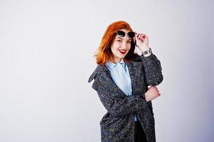 Portrait of a fantastic redheaded girl in blue shirt, grey overcoat posing with sunglasses in the studio. photo