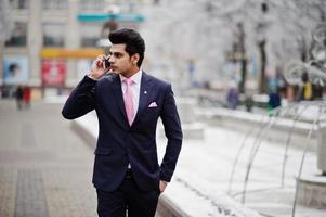 Elegant indian macho man model on suit and pink tie posed on winter day and speaking on mobile phone. photo