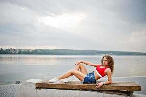 Sexy curly model girl in red top, jeans denim shorts, eyeglasses and sneakers posed on bench against lake. photo