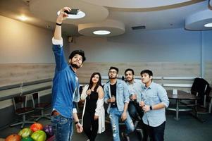 Group of five south asian peoples having rest and fun at bowling club. Making selfie by phone and holding cold soda drinks from glass bottles. photo