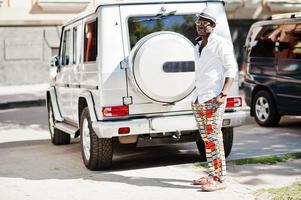 Stylish african american man in white shirt and colored pants with hat and glasses posed outdoor against silver mafia suv car. Black fashionable model boy. photo