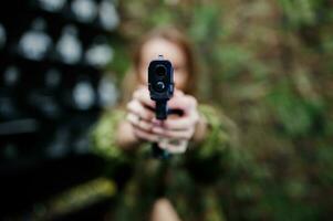 Military girl in camouflage uniform with gun at hand against army background on shooting range. Focus on gun. photo