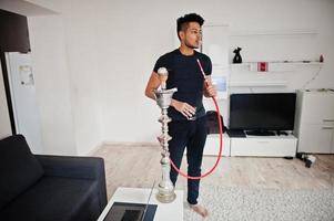 Handsome and fashionable indian man in black standing at room, smoking hookah with glasses at hand. photo