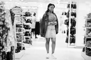 Modish african american woman in  red shirt and jeans skirt with jacket posed at clothes store. It's time for shopping. photo