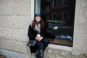 chica mexicana rizada con gorra de cuero y taza de café de plástico a mano caminando por las calles de la ciudad. foto