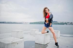 Chica modelo rizada sexy en top rojo, pantalones cortos de mezclilla, chaqueta de cuero y zapatillas de deporte posadas en cubos de piedra. foto