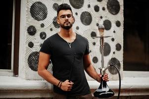 Stylish beard arabian man in glasses and black t-shirt smoking hookah outdoor. Arab model having rest. photo