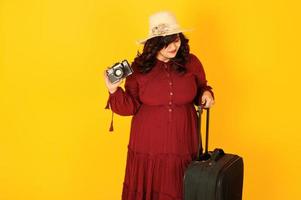 atractiva mujer viajera del sur de asia con vestido rojo intenso, sombrero posado en el estudio sobre fondo amarillo con maleta y cámara de fotos antigua.