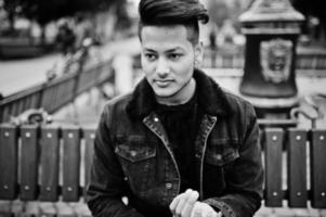 Handsome and fashionable indian man in black jeans jacket posed outdoor, sitting on bench. photo