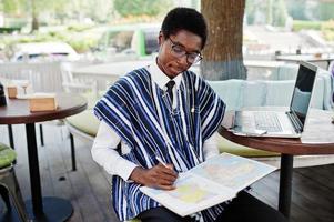 African man in traditional clothes and glasses sitting behind laptop at outdoor caffe and looking on map of Africa and Ghana at his notebook. photo