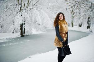 Elegance curly girl in fur coat at snowy forest park at winter. photo