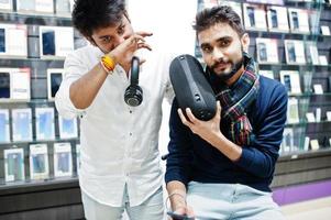 Two indians mans customer buyer at mobile phone store with earphones and wireless speaker listening music. South asian peoples and technologies concept. Cellphone shop. photo