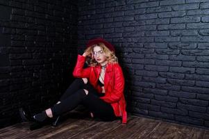 Studio portrait of blonde girl with red hat, glasses and leather jacket posed against brick wall. photo