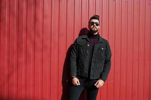 Fashion Arab man wear on black jeans jacket and sunglasses posed against red steel wall background. Stylish and fashionable arabian model guy. photo