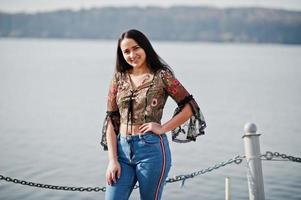 Pretty latino model girl from Ecuador wear on jeans posed against lake. photo