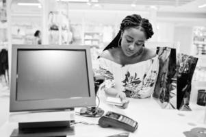 Happy african american woman customer with colored shopping bags paying by credit card near cash teminal with pos. photo