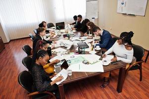 Multiracial business team addressing meeting around boardroom table, working together and write something on papers. photo