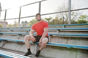 joven musculoso barbudo brutal vestido con camisa roja, pantalones cortos y gorra en el estadio. foto