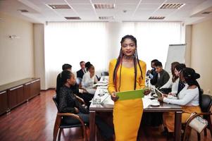 Face of handsome african business woman in yellow dress, holding clipboard on the background of business peoples multiracial team meeting, sitting in office table. photo