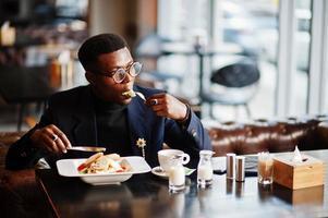 hombre afroamericano de moda con traje y gafas sentado en el café y comiendo ensalada. foto