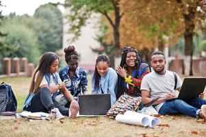 Group of five african college students spending time together on campus at university yard. Black afro friends sitting on grass and studying with laptops. photo