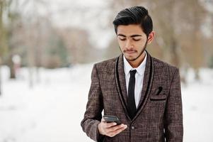 elegante estudiante indio con traje marrón y anteojos posó en el día de invierno al aire libre con el teléfono móvil a mano. foto