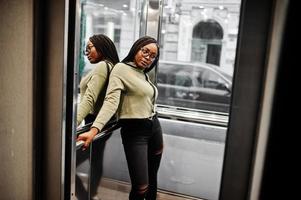 Portrait of positive young dark skinned female wearing green hoody and eyeglasses standing at elevator. photo