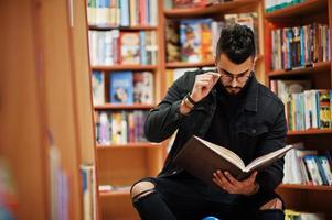 estudiante árabe alto e inteligente, vestido con chaqueta de jeans negros y anteojos, sentado en la biblioteca y leyendo un libro en las manos. foto