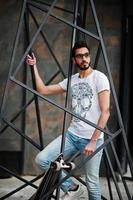 Street style arab man in eyeglasses with longboard posed inside metal pyramid construction. photo