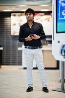 Stylish indian man posed at fast food cafe near self pay floor kiosk with mobile phone at hand. photo