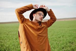 hombre elegante con gafas, chaqueta marrón y sombrero posado en campo verde. foto