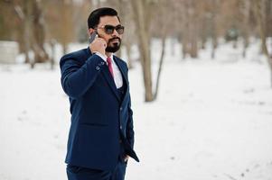 Stylish indian beard business man in suit and sunglasses posed at winter day outdoor, speaking on mobile phone. photo