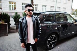 elegante hombre de barba india con chaqueta de cuero negro y gafas de sol contra un coche suv de negocios. modelo india posó al aire libre en las calles de la ciudad. foto